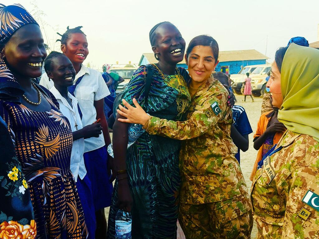 Mlinda amani wa kike wa Pakistan aliyevaa sare za kijeshi anamkumbatia mwanamke mwenye tabasamu akiwa amevaa mavazi ya kitamaduni ya Sudan Kusini, akiwa amezungukwa na wanawake wengine wa eneo hilo na walinda amani.