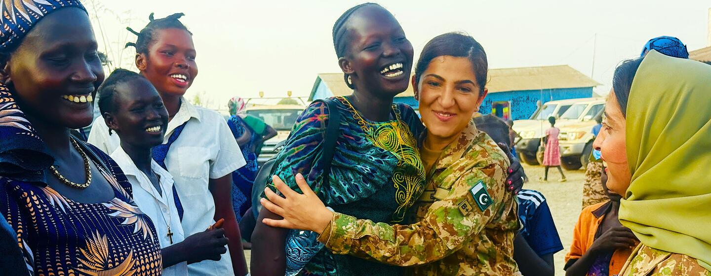 فوجی وردی میں ایک خاتون پاکستانی امن دستہ جنوبی سوڈانی روایتی لباس میں مسکراتی ہوئی خاتون کو گلے لگا رہی ہے، جسے دیگر مقامی خواتین اور امن دستوں نے گھیر رکھا ہے۔