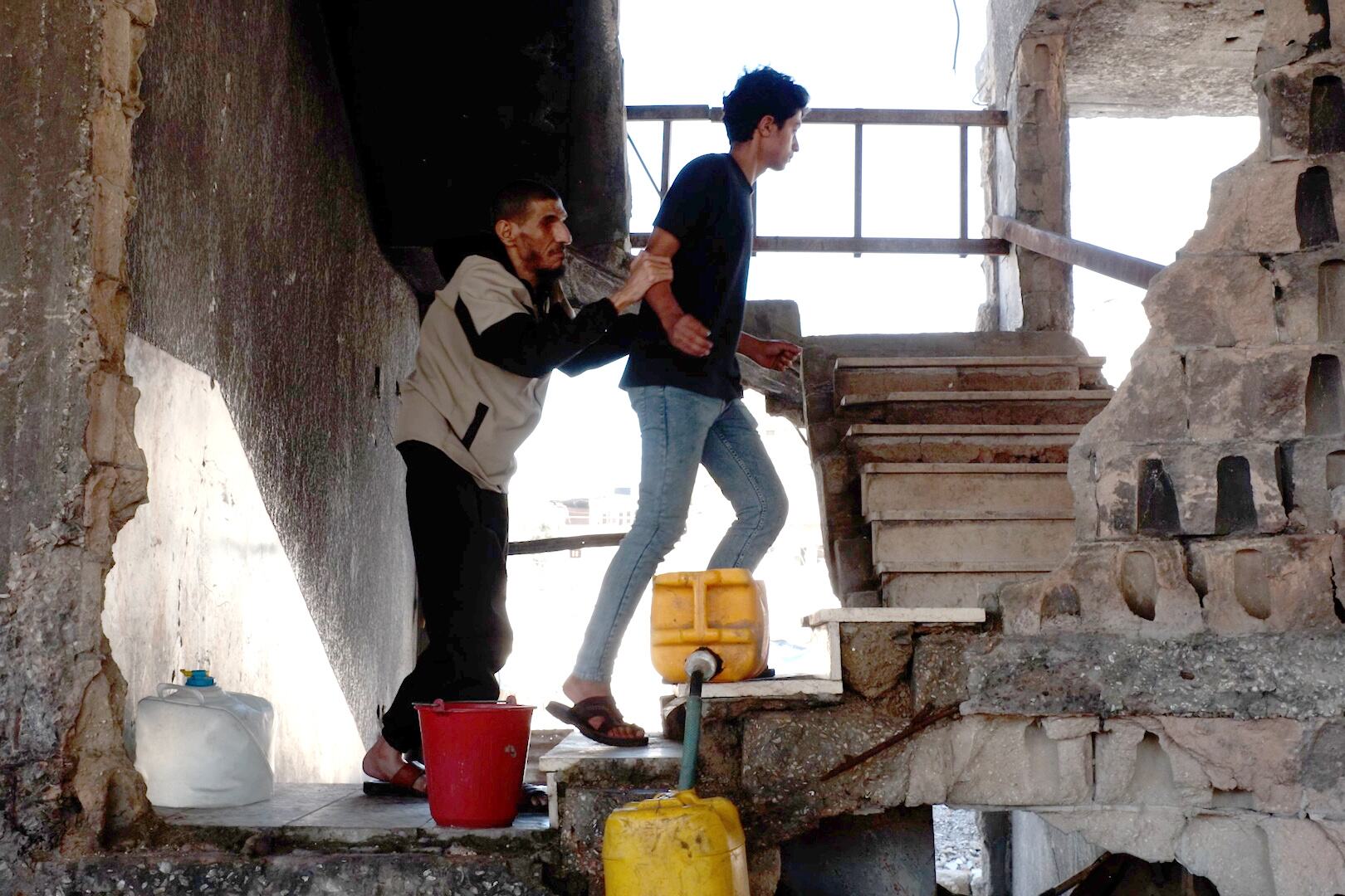 رجل أعمى يتكئ على كتف ابنه وهو يساعده على صعود مجموعة من السلالم في مبنى متضرر.