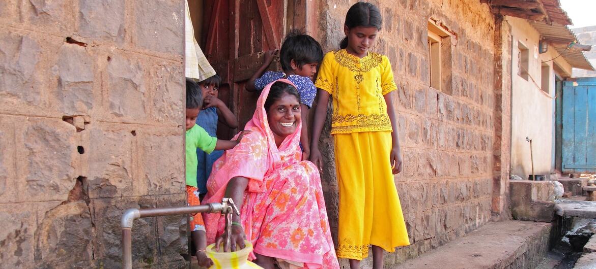 एक ग्रामीण गांव के परिवेश में, गुलाबी साड़ी पहने एक मुस्कुराती हुई भारतीय महिला धातु के नल से पीले और सफेद रंग के पानी के पात्र में पानी भर रही है, और बच्चे पास ही में यह सब देख रहे हैं।