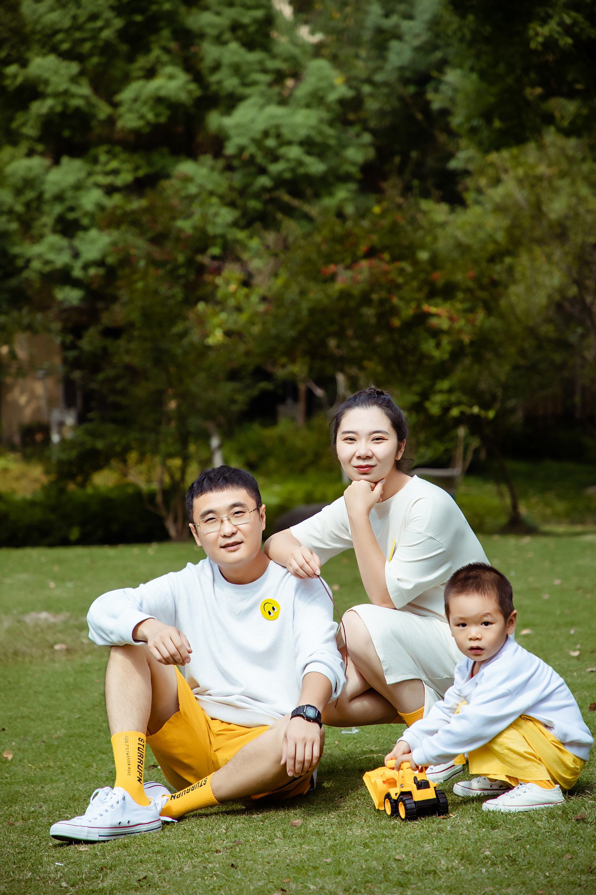 一家三口在公园的草坪上合影，父亲和母亲坐在他们的小儿子身后。