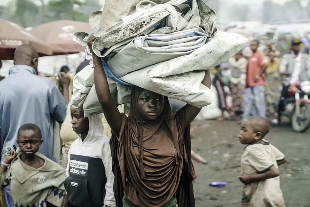 À Goma, dans un camp de réfugiés, une jeune fille africaine porte sur sa tête un gros paquet d'affaires, entourée d'autres enfants.