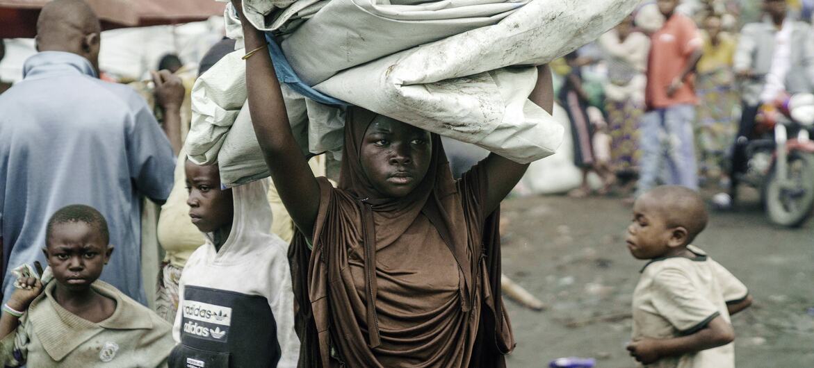 À Goma, dans un camp de réfugiés, une jeune fille africaine porte sur sa tête un gros paquet d'affaires, entourée d'autres enfants.
