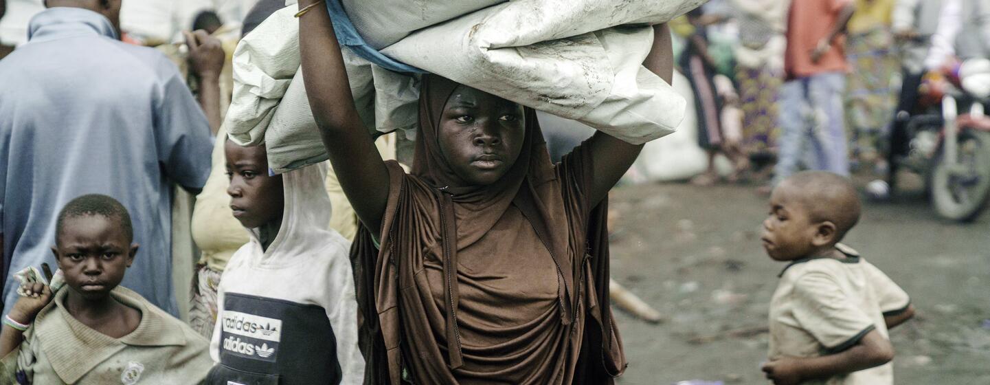 À Goma, dans un camp de réfugiés, une jeune fille africaine porte sur sa tête un gros paquet d'affaires, entourée d'autres enfants.