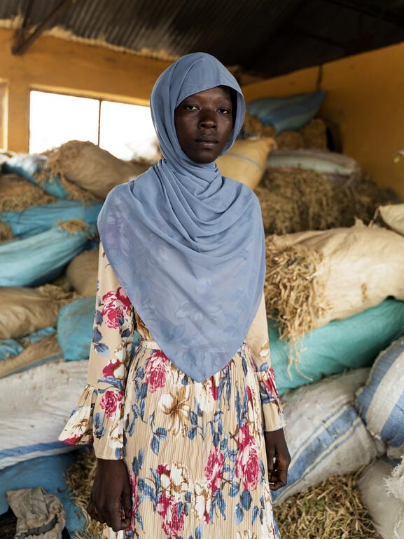 Une femme portant un hijab bleu clair et une robe à fleurs se tient à l'intérieur d'un entrepôt rempli de sacs de céréales ou de foin.