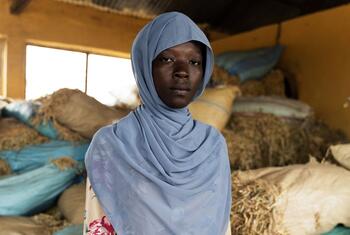 A woman wearing a light blue hijab and a floral dress stands inside a storage facility filled with sacks of grain or hay.
