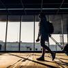 Silhouette of a woman with a backpack and rolling suitcase walking through a modern airport terminal towards a large window with bright sunlight.