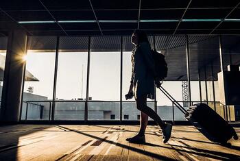 Silhouette of a woman with a backpack and rolling suitcase walking through a modern airport terminal towards a large window with bright sunlight.
