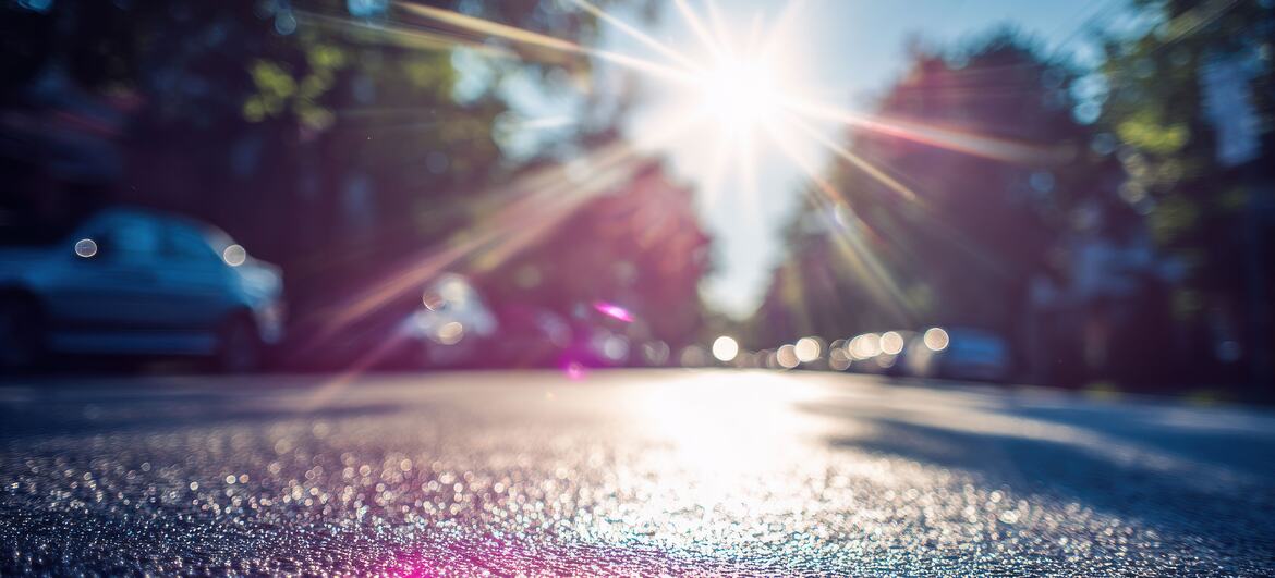 Intense sunlight beams through trees, creating a shimmering heatwave effect on wet asphalt in an urban street setting.