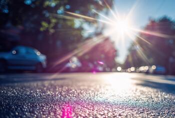 Intense sunlight beams through trees, creating a shimmering heatwave effect on wet asphalt in an urban street setting.