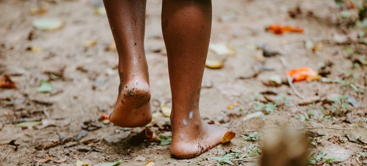 A close-up of a person's bare feet walking on a muddy forest floor covered with leaves.