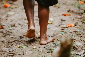 A close-up of a person's bare feet walking on a muddy forest floor covered with leaves.