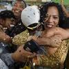 Une femme vêtue d'un chemisier à motifs dorés enlace un homme coiffé d'une casquette blanche lors d'une fête, entourée de monde et sous une pluie de confettis.