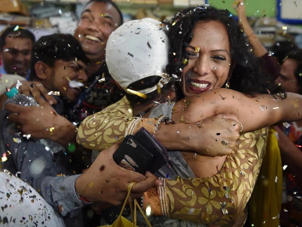 Une femme vêtue d'un chemisier à motifs dorés enlace un homme coiffé d'une casquette blanche lors d'une fête, entourée de monde et sous une pluie de confettis.