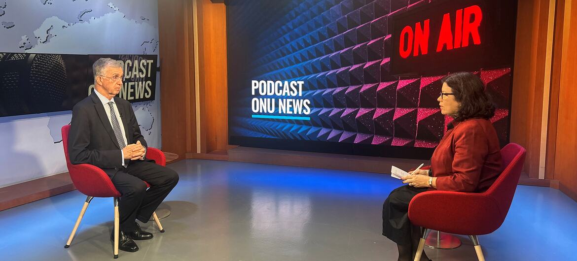Um homem e uma mulher estão sentados em um estúdio de televisão para uma entrevista. Uma grande tela atrás deles exibe os textos "NO AR" e "PODCAST ONU NEWS".