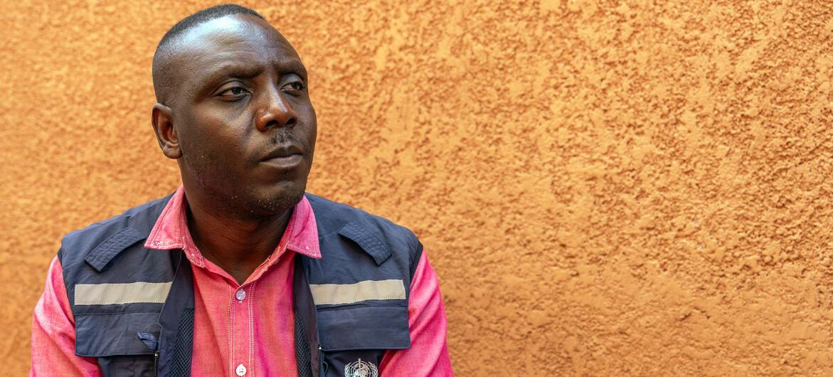 Le Dr Chris Opesen, anthropologue à l'Organisation mondiale de la santé en Ouganda, regarde pensivement sur le côté, vêtu d'une chemise rose et d'un gilet bleu marine orné du logo de l'OMS, devant un mur orange texturé.