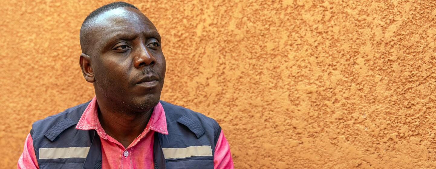 Dr Chris Opesen, an anthropologist with the World Health Organization in Uganda, looks thoughtfully to the side while wearing a pink shirt and a navy vest with the WHO logo, against a textured orange wall.