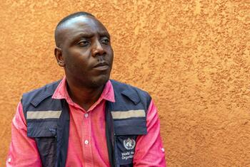 Le Dr Chris Opesen, anthropologue à l'Organisation mondiale de la santé en Ouganda, regarde pensivement sur le côté, vêtu d'une chemise rose et d'un gilet bleu marine orné du logo de l'OMS, devant un mur orange texturé.