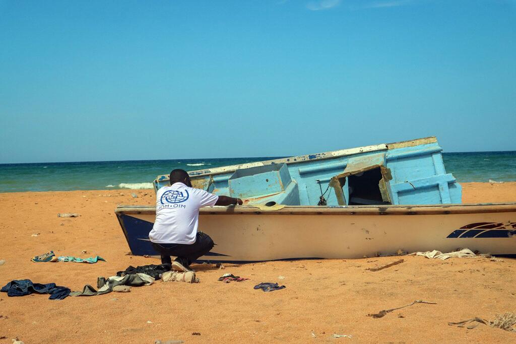 Mwanamume aliyevaa shati jeupe la IOM amejilaza kwenye ufuo wa mchanga karibu na mashua ya bluu na nyeupe iliyoharibika, huku bahari ikiwa nyuma.