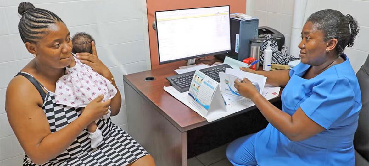 Una enfermera con uniforme azul atiende a una madre que sostiene a su bebé recién nacido en una clínica. La madre está sentada mientras la enfermera revisa documentos en un escritorio con una computadora.
