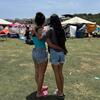 Dos mujeres permanecen abrazadas en un campamento de ayuda humanitaria al aire libre, con tiendas de campaña y suministros al fondo.