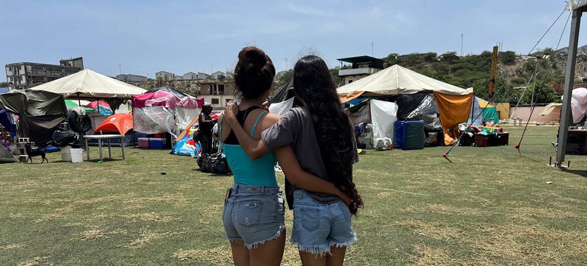 Dos mujeres permanecen abrazadas en un campamento de ayuda humanitaria al aire libre, con tiendas de campaña y suministros al fondo.