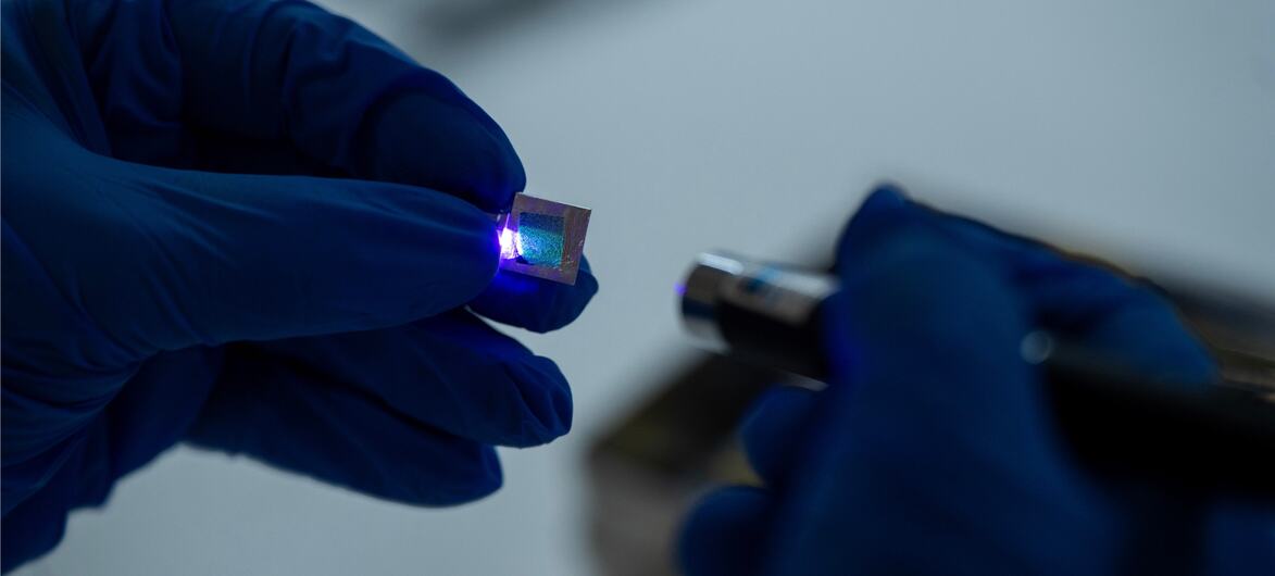 Um cientista com luvas azuis segura um pequeno microchip quadrado sob uma luz azul para inspeção em um ambiente de laboratório.