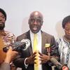 Mwanamume aliyevaa suti akizungumza kwenye maikrofoni wakati wa hafla ya Siku ya Kiswahili, akiwa amezungukwa na watu wengine wawili.