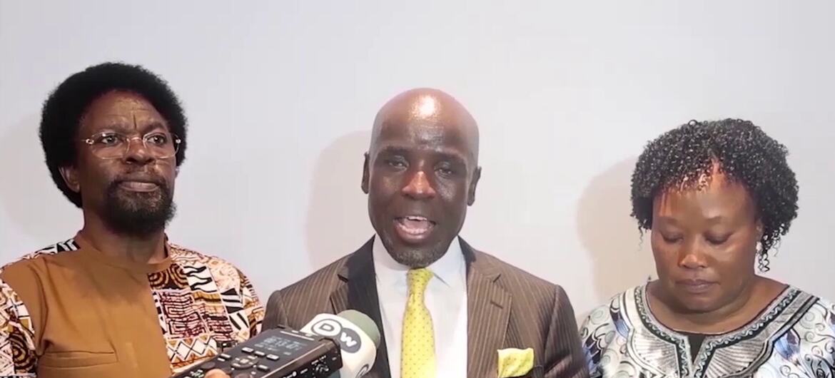 Mwanamume aliyevaa suti akizungumza kwenye maikrofoni wakati wa hafla ya Siku ya Kiswahili, akiwa amezungukwa na watu wengine wawili.