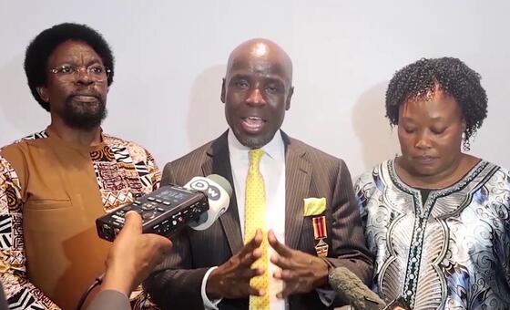 Mwanamume aliyevaa suti akizungumza kwenye maikrofoni wakati wa hafla ya Siku ya Kiswahili, akiwa amezungukwa na watu wengine wawili.