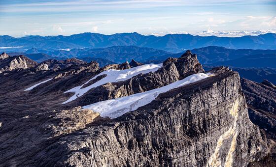 A view of the snow-capped peaks and glaciers of a mountain range in Papua, Indonesia, with distant blue mountains under a clear sky.