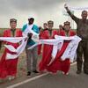 Группа людей, в том числе две женщины в традиционной красно-золотой одежде, стоят на обочине дороги, держа в руках длинный белый пояс. Мужчина в камуфляжной рубашке поднимает руки, а женщина слева держит фотоаппарат.