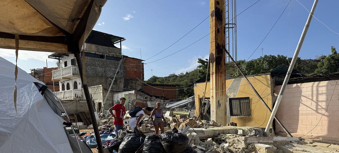 Varias personas permanecen de pie entre los escombros y una tienda de campaña blanca en una zona afectada por un desastre en Venezuela.