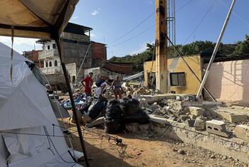 Varias personas permanecen de pie entre los escombros y una tienda de campaña blanca en una zona afectada por un desastre en Venezuela.