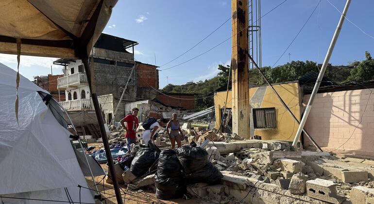 Varias personas permanecen de pie entre los escombros y una tienda de campaña blanca en una zona afectada por un desastre en Venezuela.