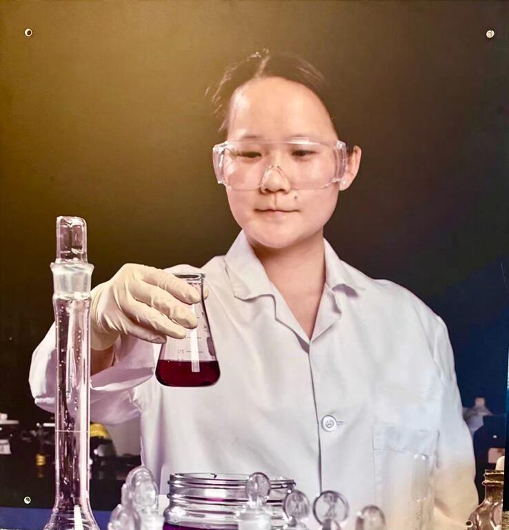 一位身穿实验服、戴着护目镜的亚裔女科学家正在实验室里检查一个装有红色液体的烧杯。