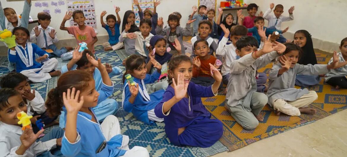 پاکستانی سکول کے بچوں کا ایک گروپ کلاس روم میں ہاتھ اٹھا کر مسکرا رہا ہے۔ وہ ایک نمونہ دار قالین پر بیٹھے ہیں، سیکھنے کا سامان پکڑے ہوئے ہیں۔
