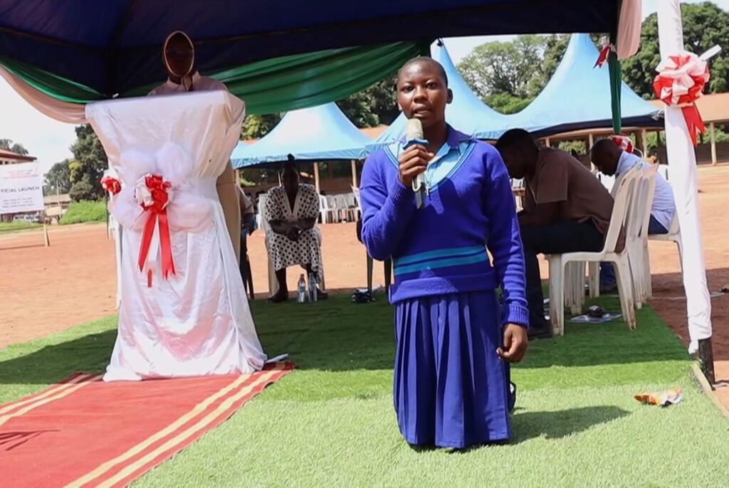Mwanamke kijana aliyevaa sare ya shule ya bluu akizungumza kwenye kipaza sauti katika hafla iliyo chini ya hema lililopambwa kwenye zulia jekundu. Wahudhuriaji wengine wameketi nyuma kwenye uwanja wa vumbi.