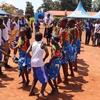 Kundi la watoto na vijana waliovalia mavazi ya rangi mbalimbali wakicheza densi katika hafla ya nje huku mahema ya bluu yakiwa nyuma.