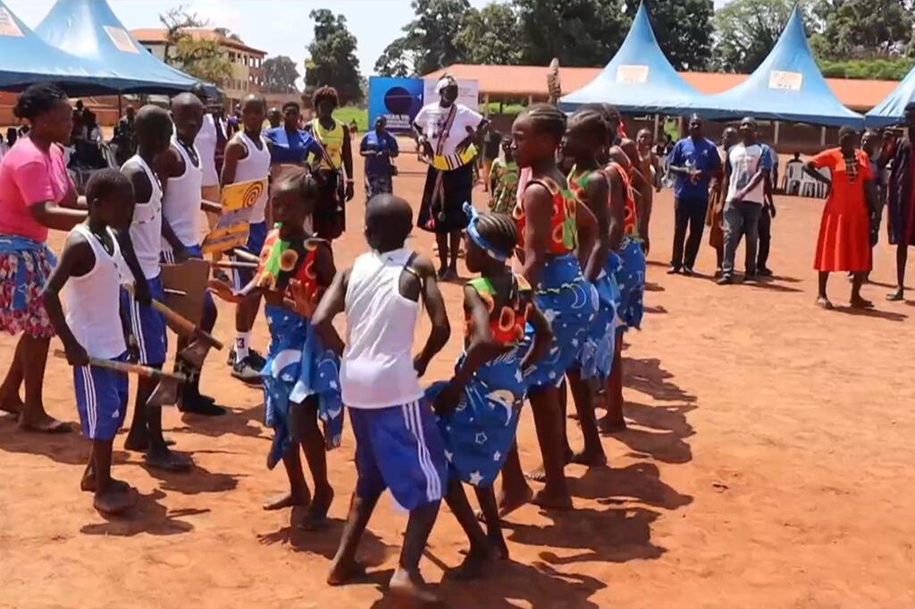 Kundi la watoto na vijana waliovalia mavazi ya rangi mbalimbali wakicheza densi katika hafla ya nje huku mahema ya bluu yakiwa nyuma.