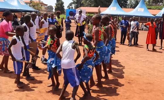 Kundi la watoto na vijana waliovalia mavazi ya rangi mbalimbali wakicheza densi katika hafla ya nje huku mahema ya bluu yakiwa nyuma.