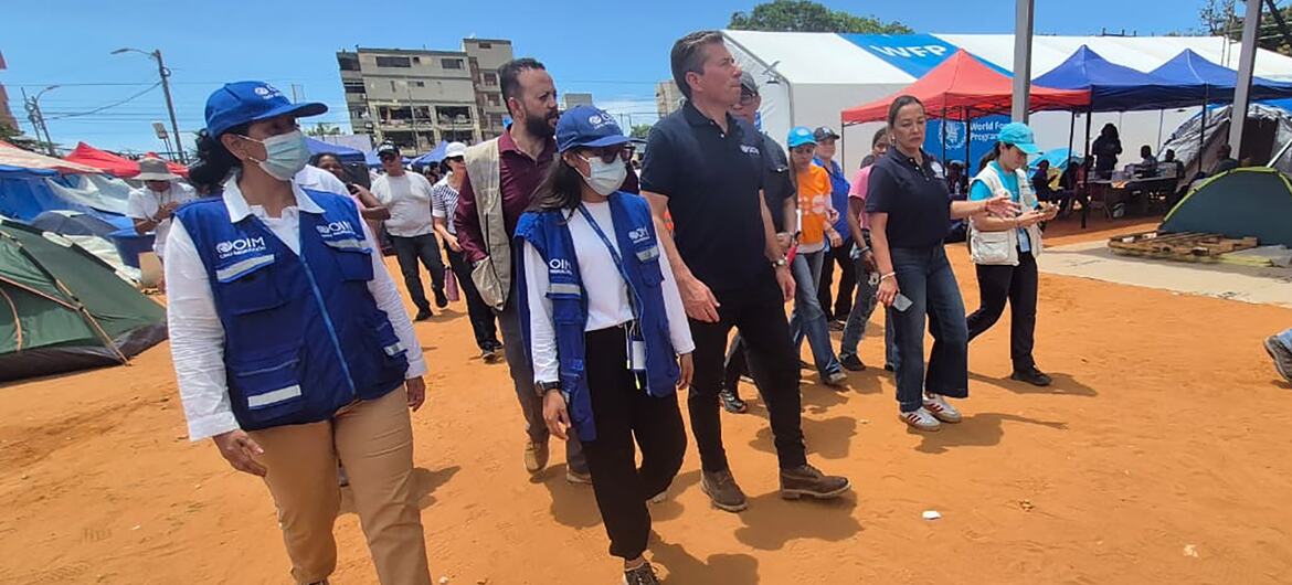 Funcionarios de la ONU y trabajadores humanitarios con chalecos azules caminan por un campo de refugiados con tiendas de campaña al fondo.