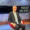 Um homem de terno está sentado em uma cadeira vermelha em um estúdio com uma placa escrito 'PODCAST ONU NEWS' ao fundo.