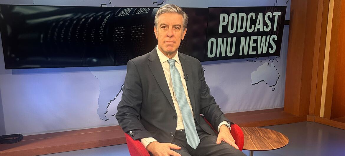 Um homem de terno está sentado em uma cadeira vermelha em um estúdio com uma placa escrito 'PODCAST ONU NEWS' ao fundo.