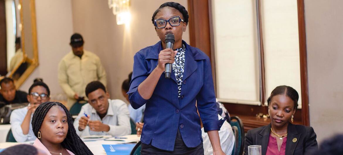 Uma mulher negra de blazer azul fala ao microfone durante a Escola de Verão Anticorrupção em Porto Príncipe, Haiti. Outros alunos estão sentados às mesas ao fundo.