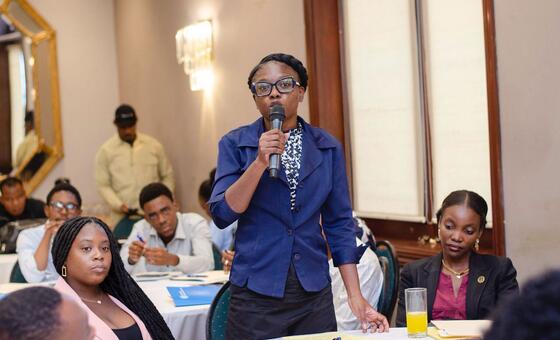 Une femme noire vêtue d'un blazer bleu prend la parole au micro lors de l'école d'été anticorruption à Port-au-Prince, en Haïti. D'autres étudiants sont assis à des tables en arrière-plan.