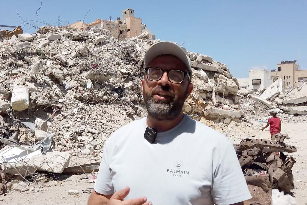 Man identified as Ramez Nabhan stands in front of a large pile of rubble in Gaza, with a microphone attached to his shirt.