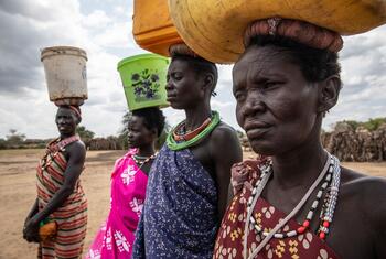 South Sudanese women carrying water containers on their heads in a rural, arid landscape.
