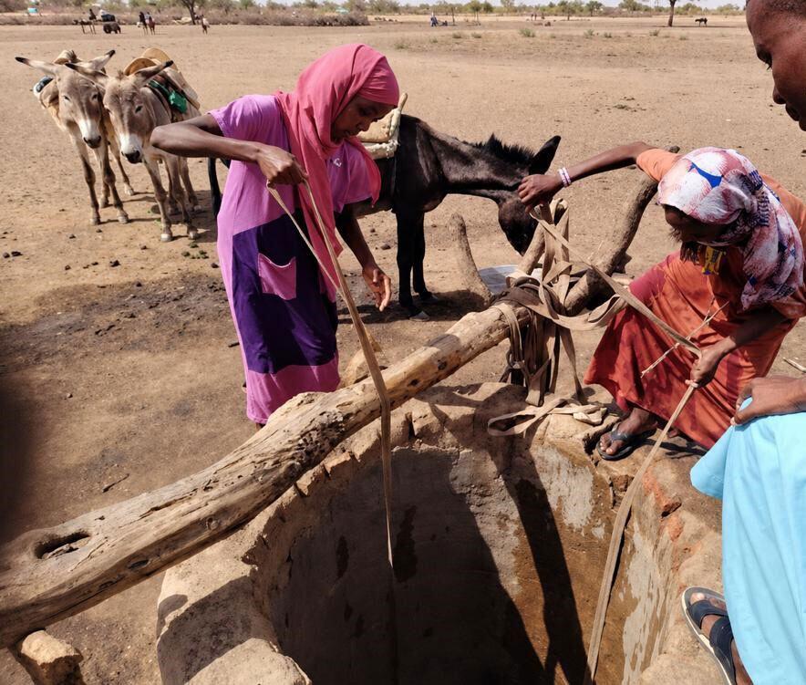 Deux femmes près d'un puits dans un paysage aride, avec des ânes à proximité.