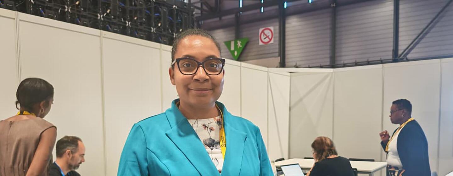 Una mujer con chaqueta azul y gafas se encuentra de pie en un evento de una conferencia, con otros asistentes al fondo.